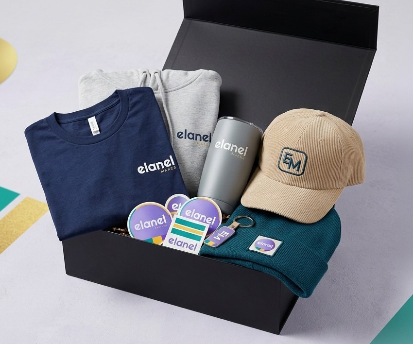 Open black gift box holding a navy tee, grey hoodie, tumbler, corduroy cap, beanie and stickers, all with the Elanel logo
