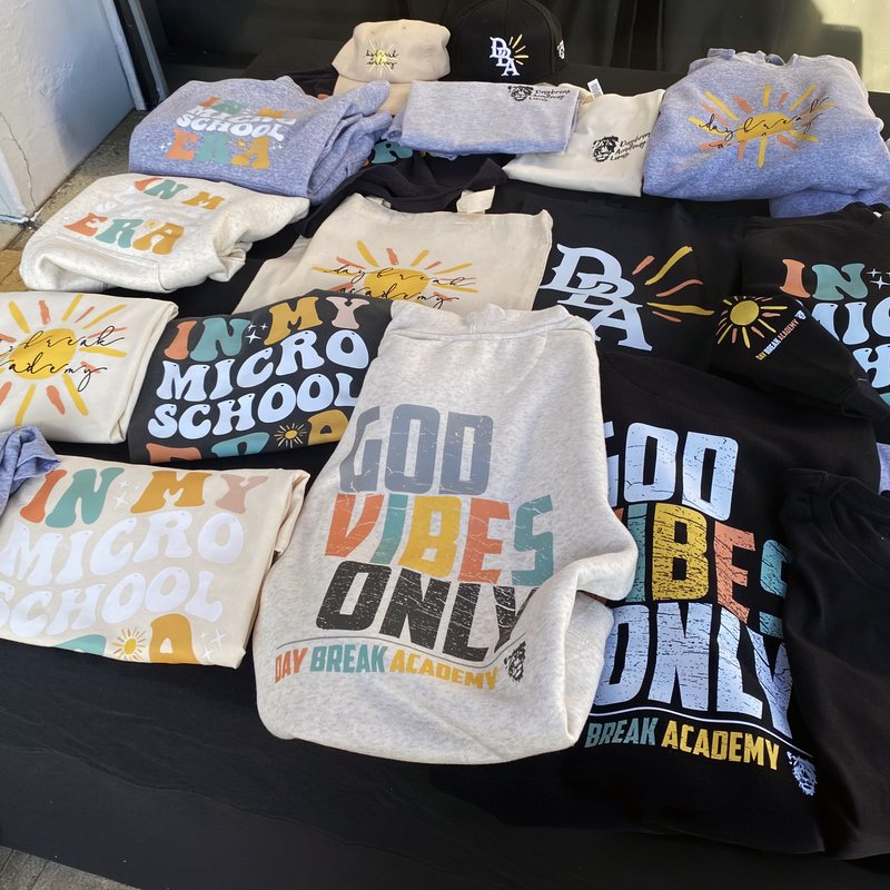 Table of finished school merchandise: printed hoodies, tees, totes and hats in several colours